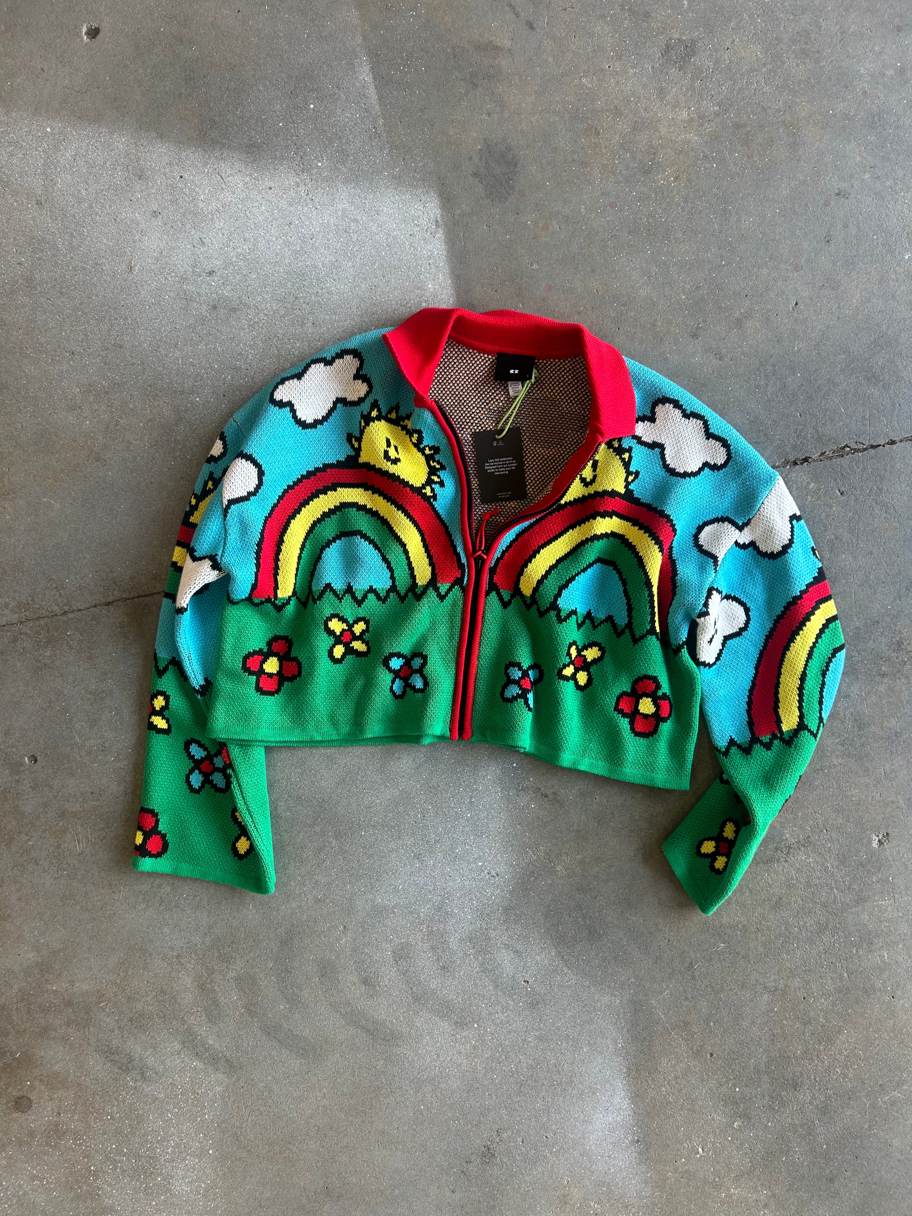 A cropped knit zip-up cardigan with a colorful cartoon landscape design, featuring rainbows, clouds, flowers, and smiling faces on a turquoise and green background, laid flat on a gray concrete floor
