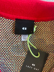 Close-up of a knit garment showing a bright red ribbed collar, a multicolor knit body with small repeating heart-like motifs, a black brand tag reading LC, a size tag, and a hanging black product tag reading LAZY OAF