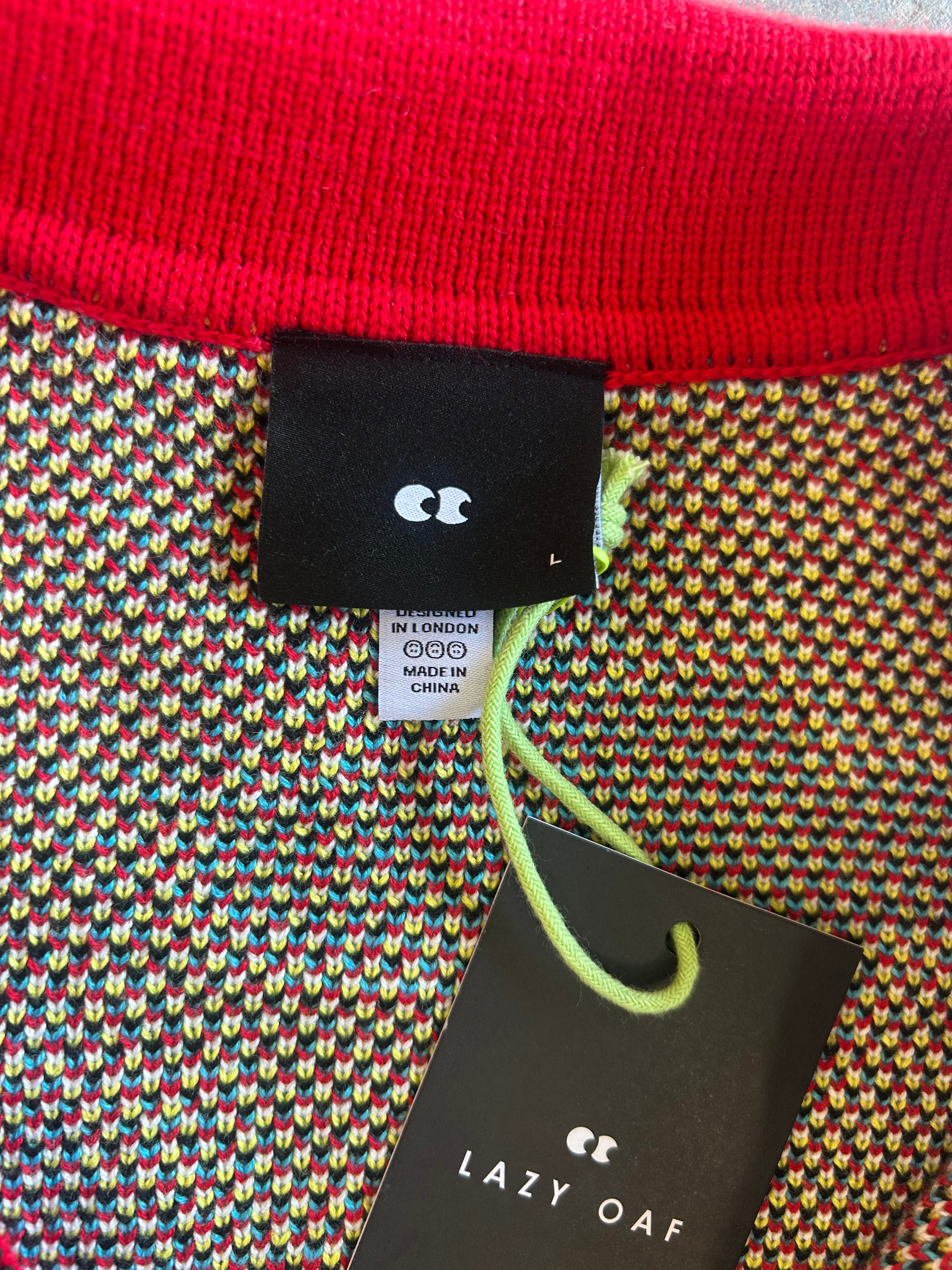 Close-up of a knit garment showing a bright red ribbed collar, a multicolor knit body with small repeating heart-like motifs, a black brand tag reading LC, a size tag, and a hanging black product tag reading LAZY OAF