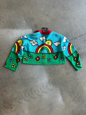 A cropped long-sleeve knit sweater with a colorful cartoon landscape print, featuring a turquoise sky, green grass, a rainbow, clouds, flowers, and a smiling sun, laid flat on a concrete floor