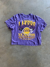 A purple Los Angeles Lakers graphic t-shirt laid flat on a gray concrete floor. The shirt features a large front print with 'LAKERS' in yellow and purple, the Lakers logo, and 'LOS ANGELES' text