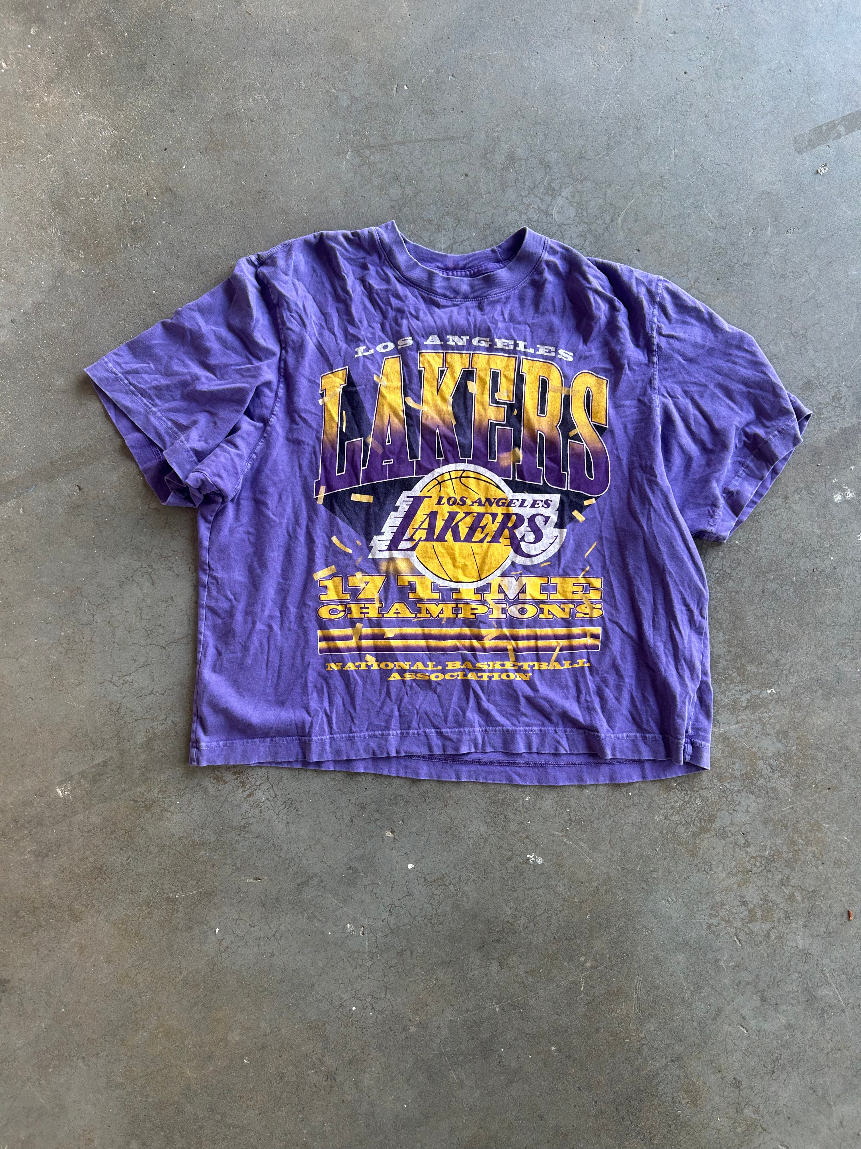 A purple Los Angeles Lakers graphic t-shirt laid flat on a gray concrete floor. The shirt features a large front print with 'LAKERS' in yellow and purple, the Lakers logo, and 'LOS ANGELES' text