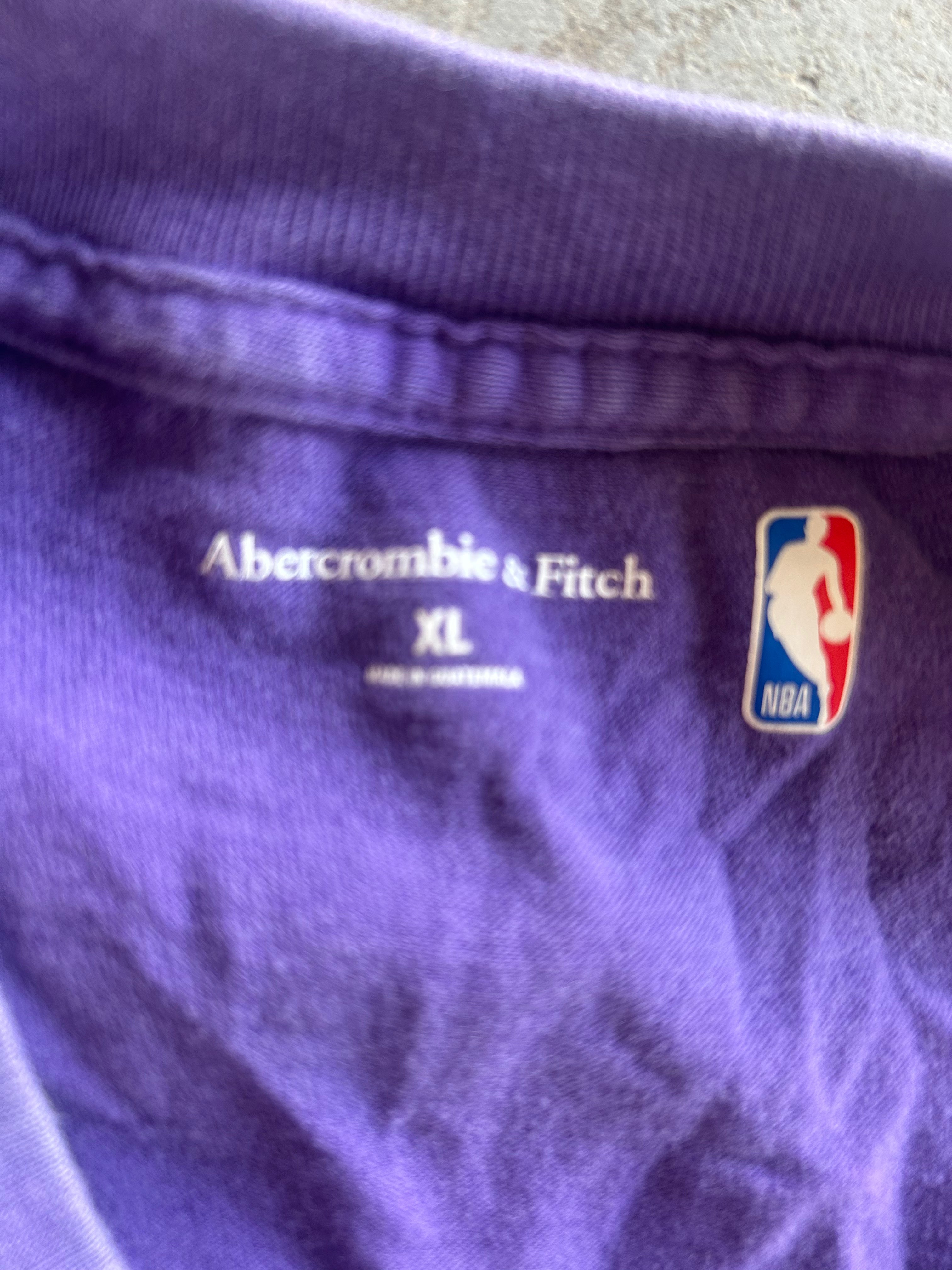 Close-up of the inside collar of a purple Abercrombie & Fitch t-shirt, showing the white brand name, size 'XL', and an NBA logo patch