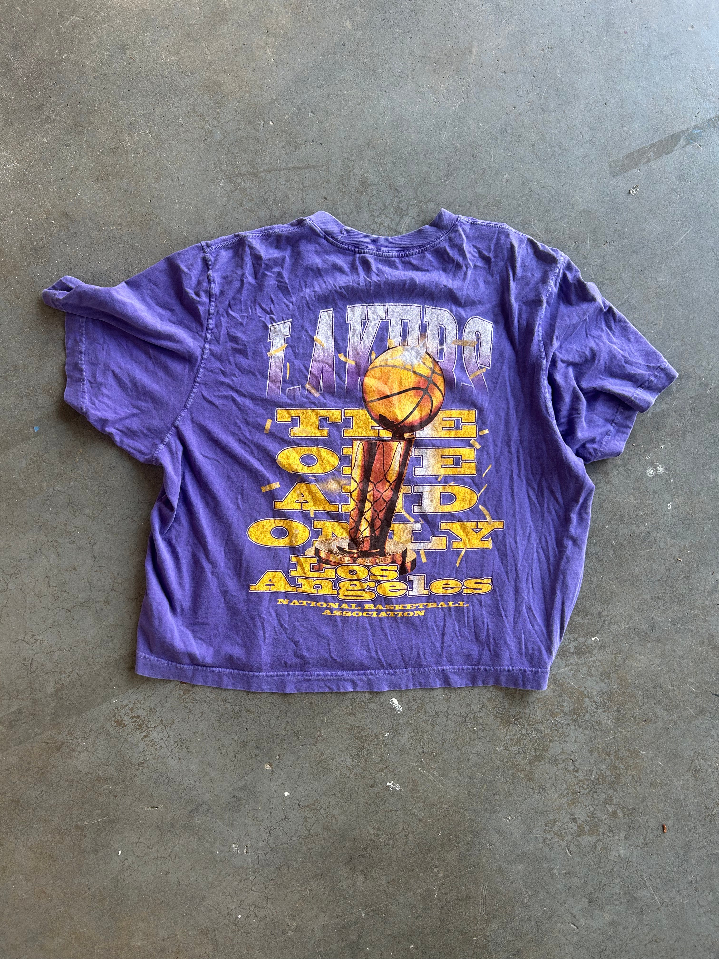 A purple Los Angeles Lakers graphic t-shirt laid flat on a gray concrete floor. The back of the shirt features a large yellow and white print with the Lakers logo and a basketball graphic