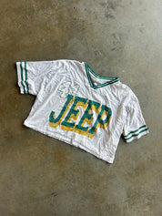 A vintage white short-sleeve Jeep jersey with green and yellow lettering and green striped sleeve cuffs, laid flat on a textured concrete floor
