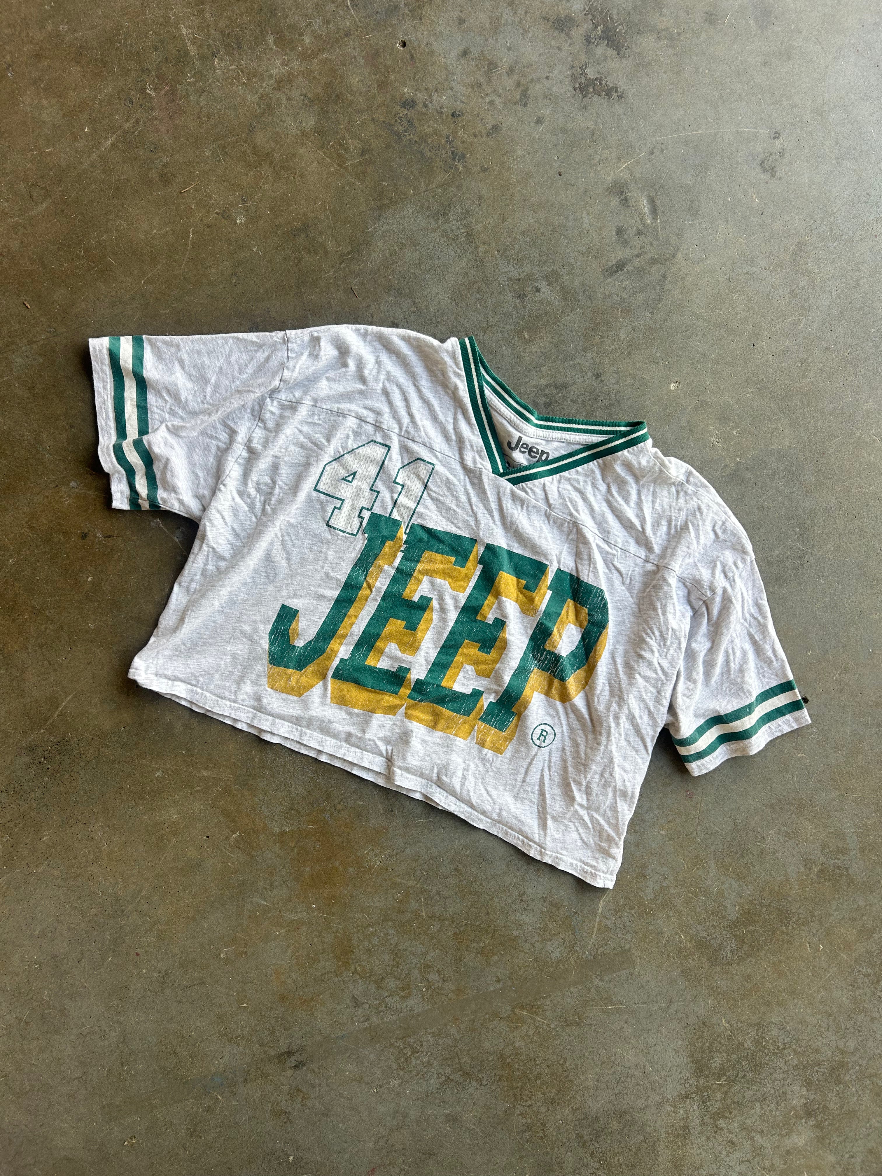 A vintage white short-sleeve Jeep jersey with green and yellow lettering and green striped sleeve cuffs, laid flat on a textured concrete floor
