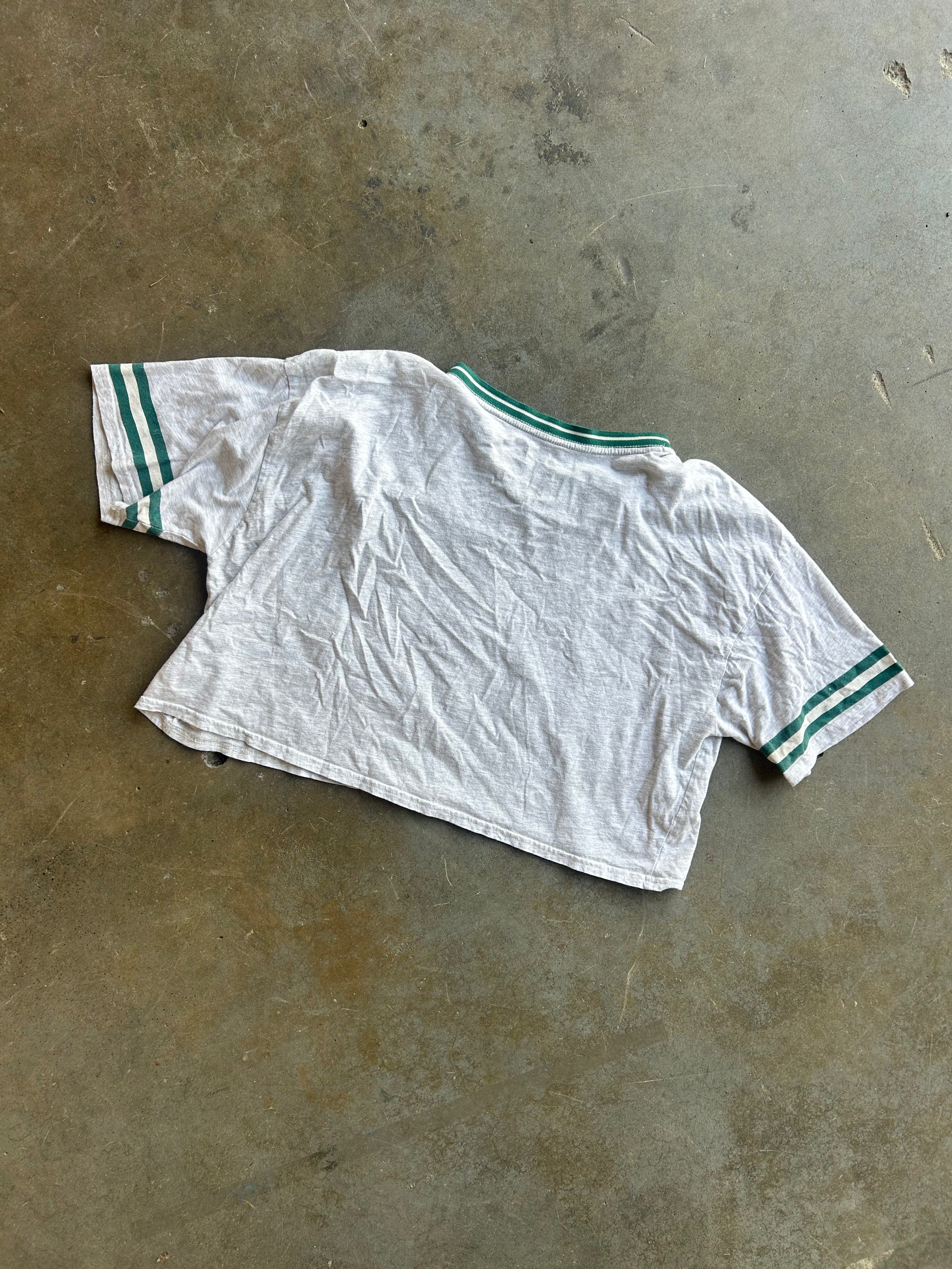 A white short-sleeve cropped T-shirt with green stripes on the collar and cuffs, laid flat on a textured concrete floor