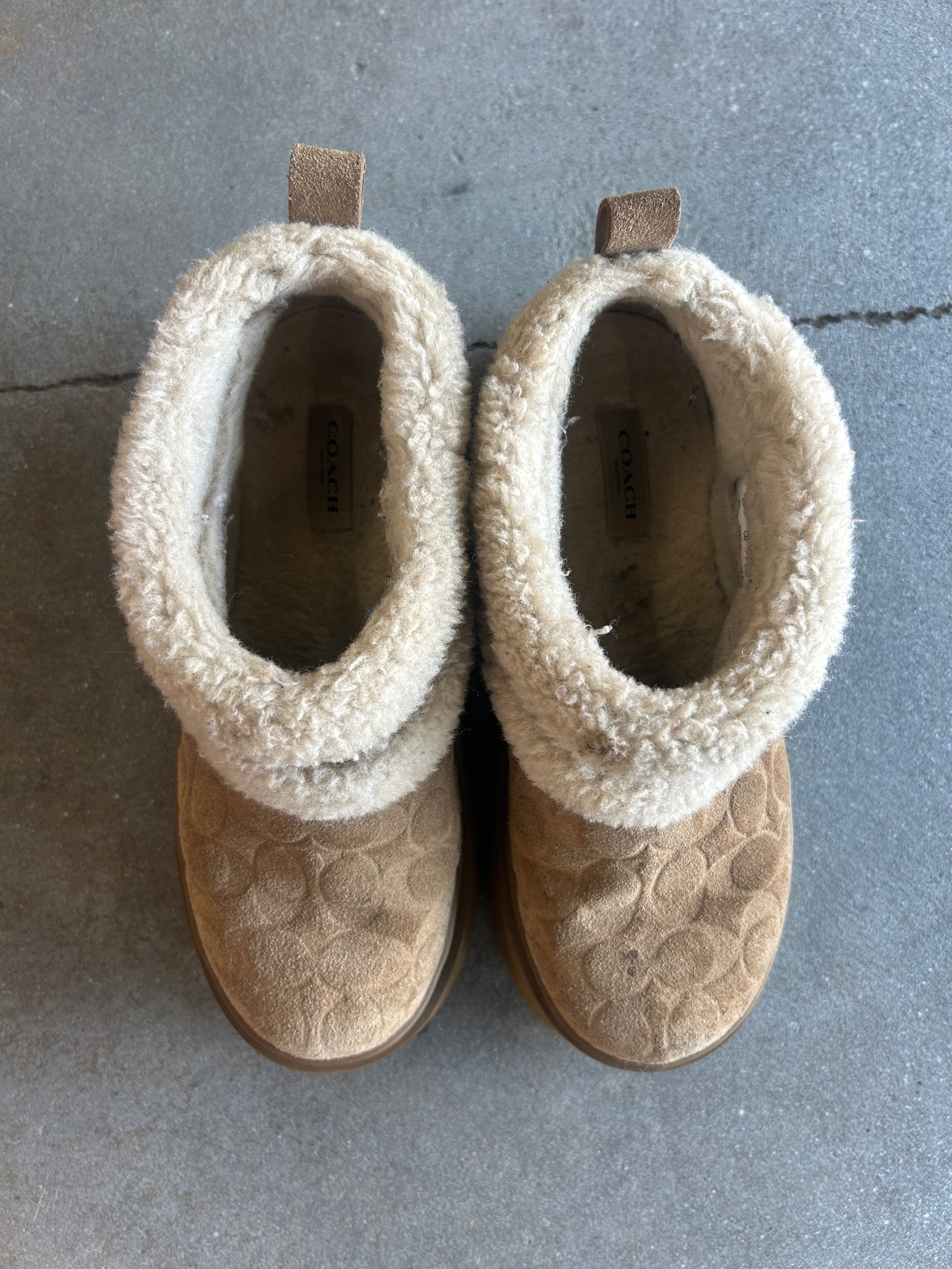 A pair of Coach ankle boots in tan with a textured chevron pattern and a thick cream sherpa collar, shown from a top-down view on a gray concrete floor