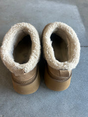 A pair of beige suede ankle boots with thick cream sherpa fleece lining, shown from a top-down view on a gray concrete floor