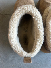 A close-up top-down view of a Coach ankle boot with a tan suede exterior and a thick cream sherpa fleece lining, showing the interior pull tab with the Coach logo