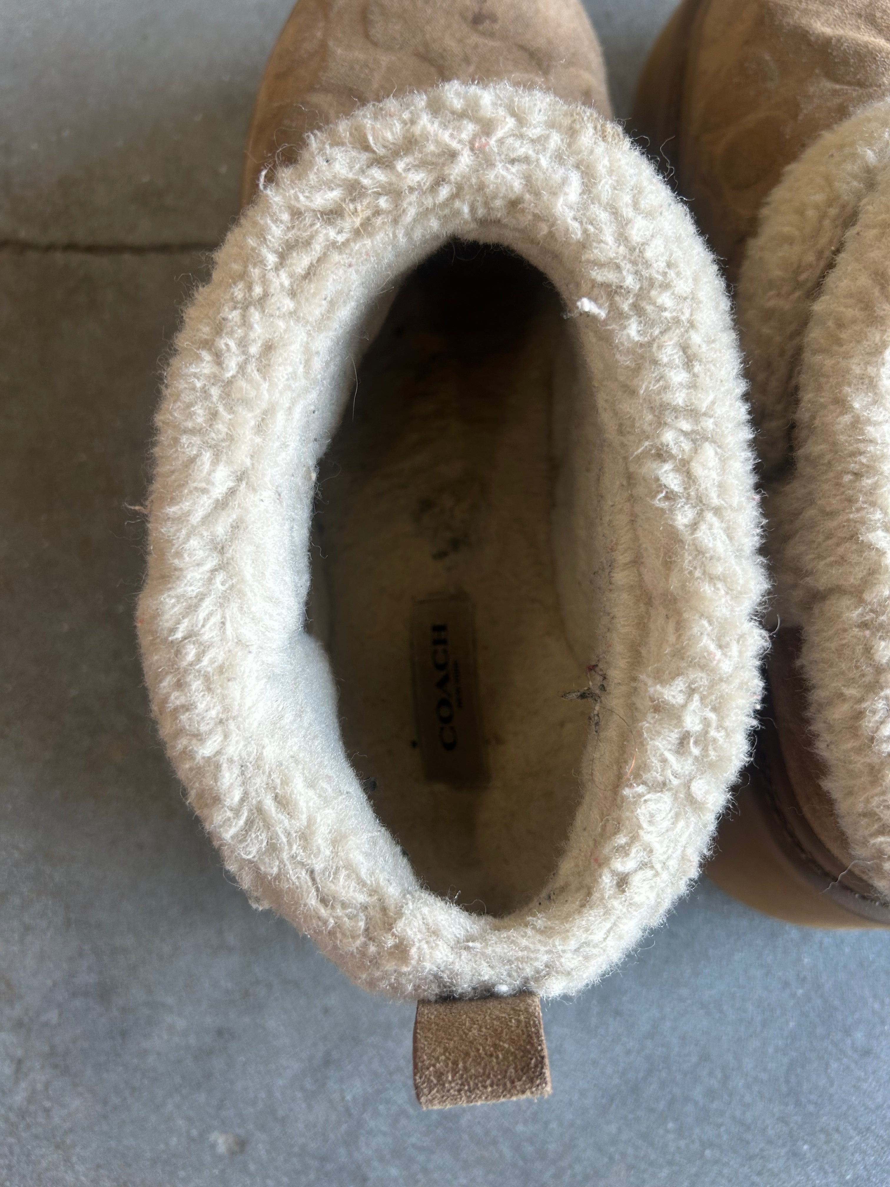 A close-up top-down view of a Coach ankle boot with a tan suede exterior and a thick cream sherpa fleece lining, showing the interior pull tab with the Coach logo