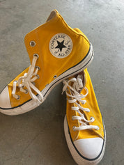 A pair of yellow Converse All Star high-top sneakers with white laces, white rubber toe caps, and a black stripe along the sole, resting on a gray concrete floor