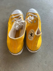 A pair of yellow Converse All Star high-top sneakers with white laces and white rubber toe caps, shown from a top-down view on a gray concrete floor