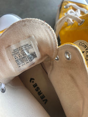 Close-up of a Converse high-top sneaker showing the beige canvas tongue label with size and country of origin details, alongside part of the yellow rubber toe cap and white laces