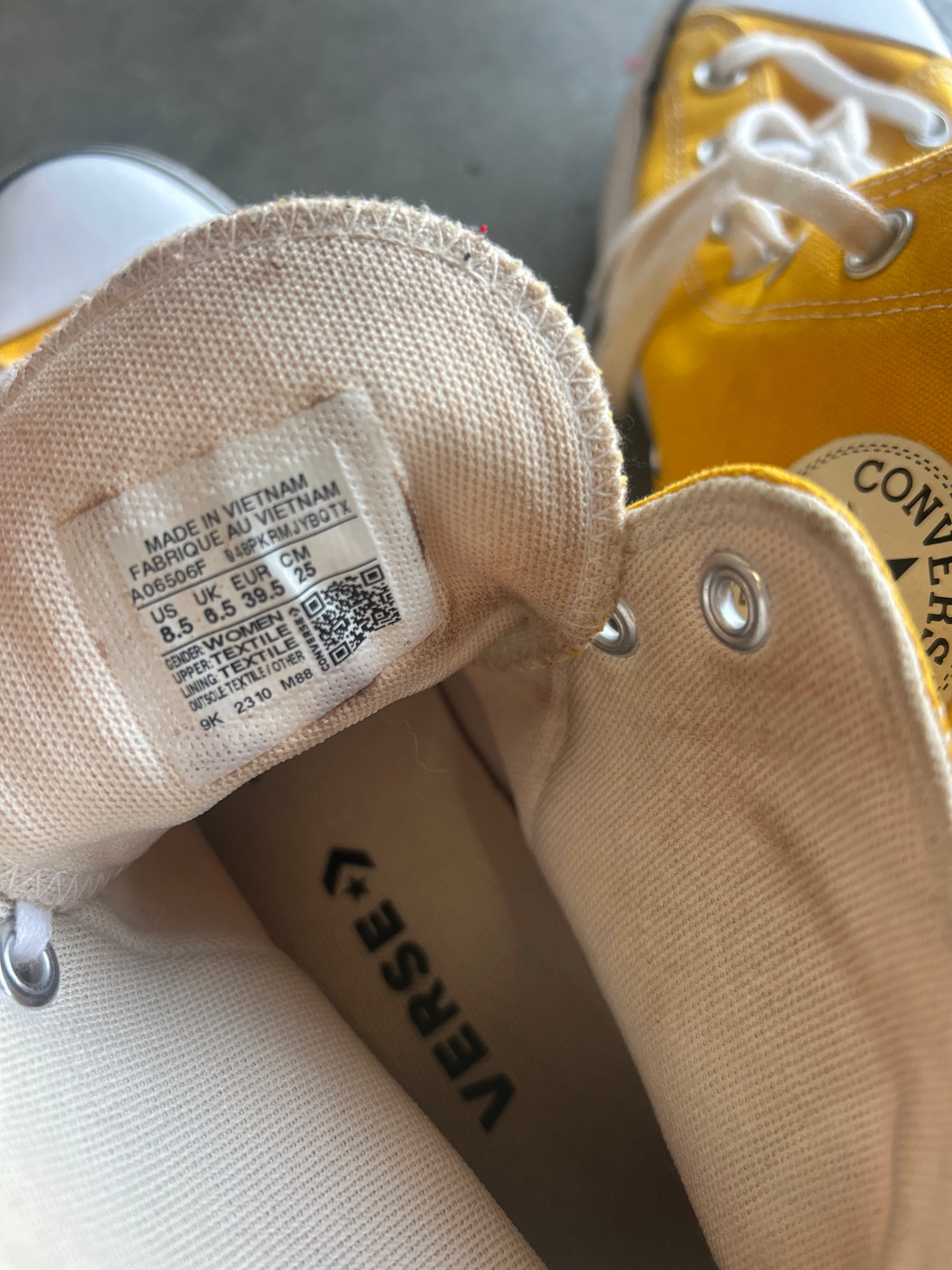 Close-up of a Converse high-top sneaker showing the beige canvas tongue label with size and country of origin details, alongside part of the yellow rubber toe cap and white laces