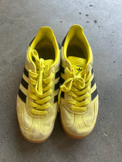 A pair of yellow Adidas Gazelle sneakers with black three-stripes and yellow laces, resting on a gray concrete floor