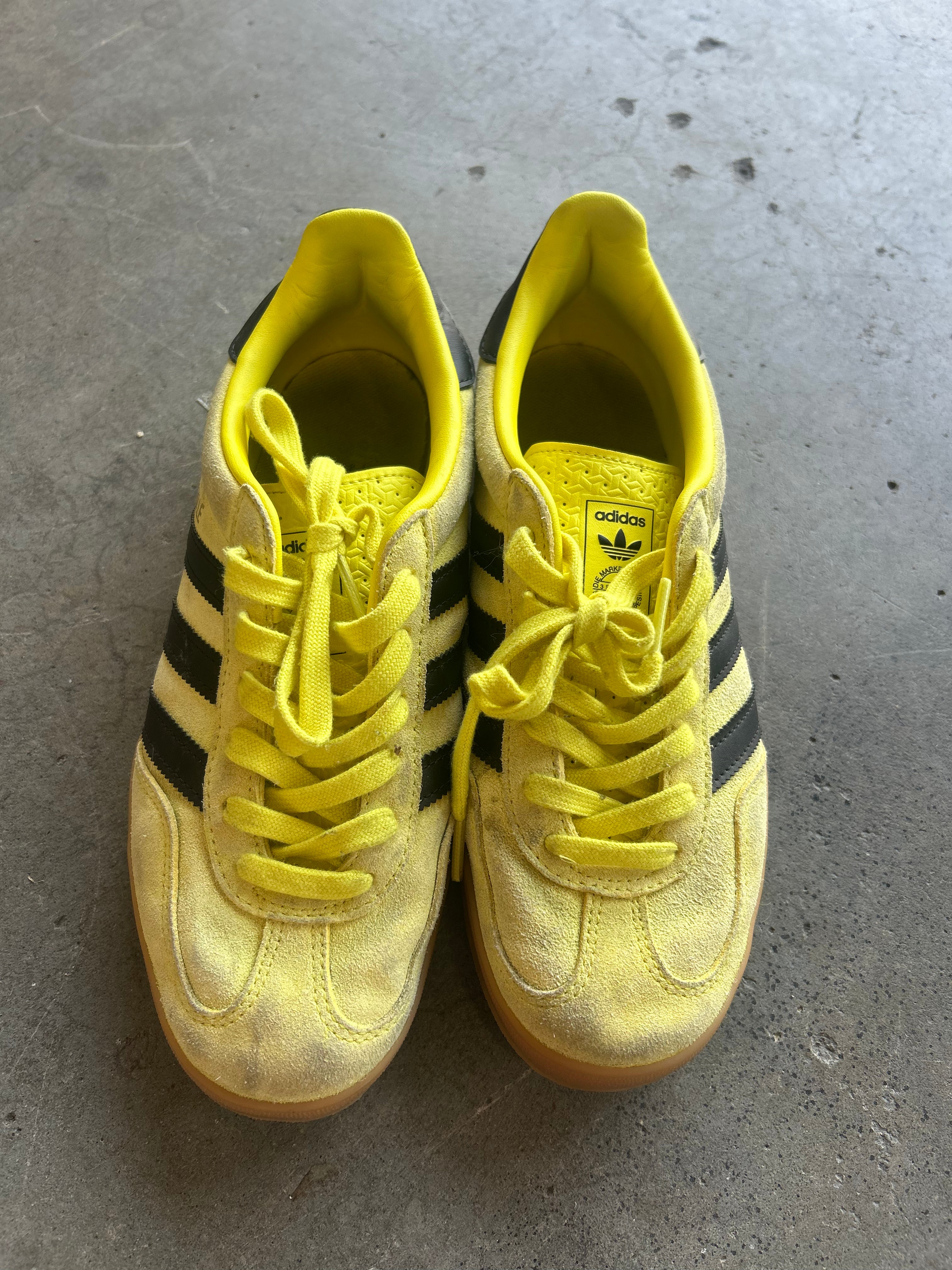 A pair of yellow Adidas Gazelle sneakers with black three-stripes and yellow laces, resting on a gray concrete floor