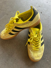 A pair of yellow Adidas Gazelle sneakers with black stripes and yellow laces, resting on a gray concrete floor