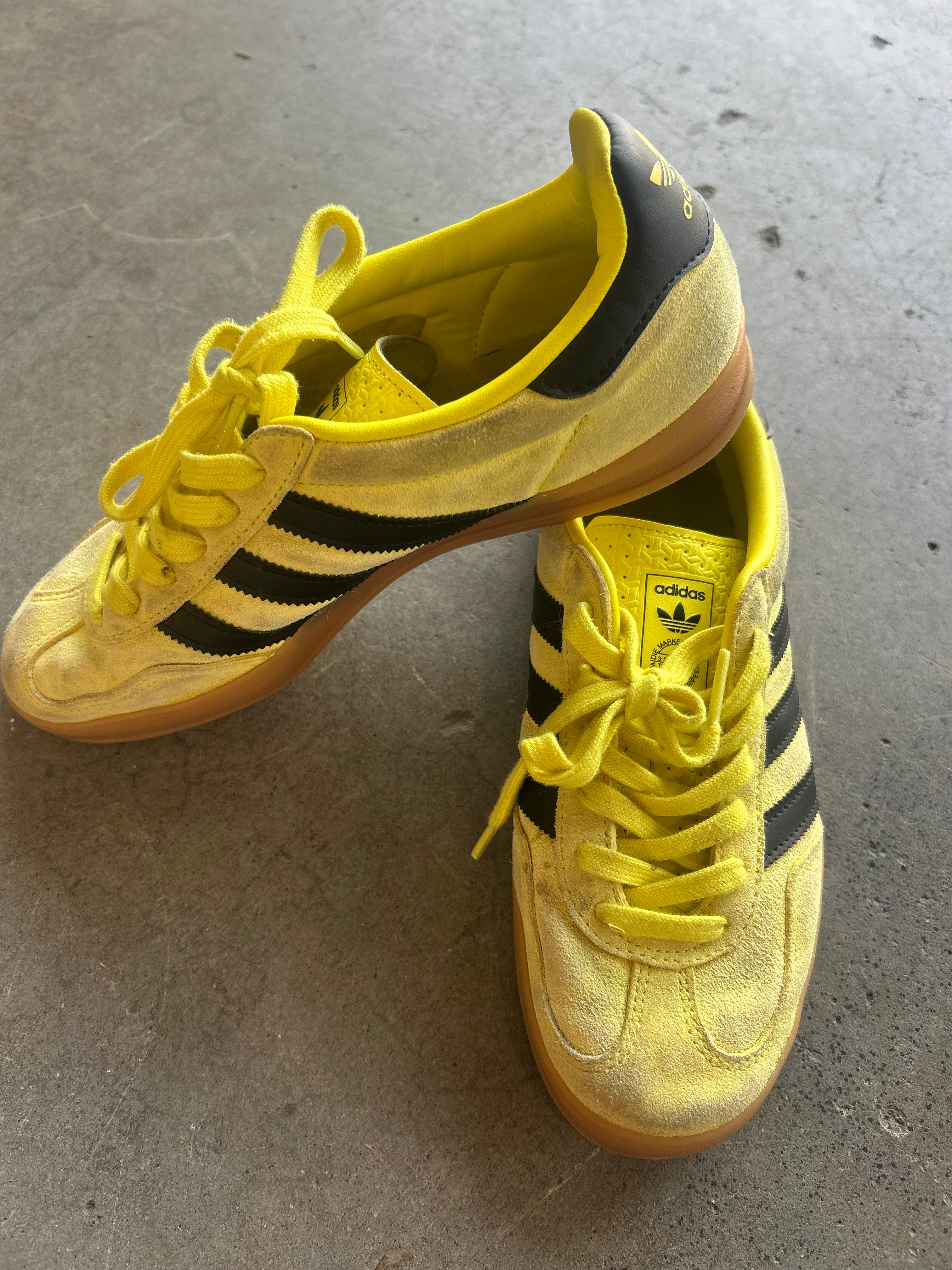 A pair of yellow Adidas Gazelle sneakers with black stripes and yellow laces, resting on a gray concrete floor