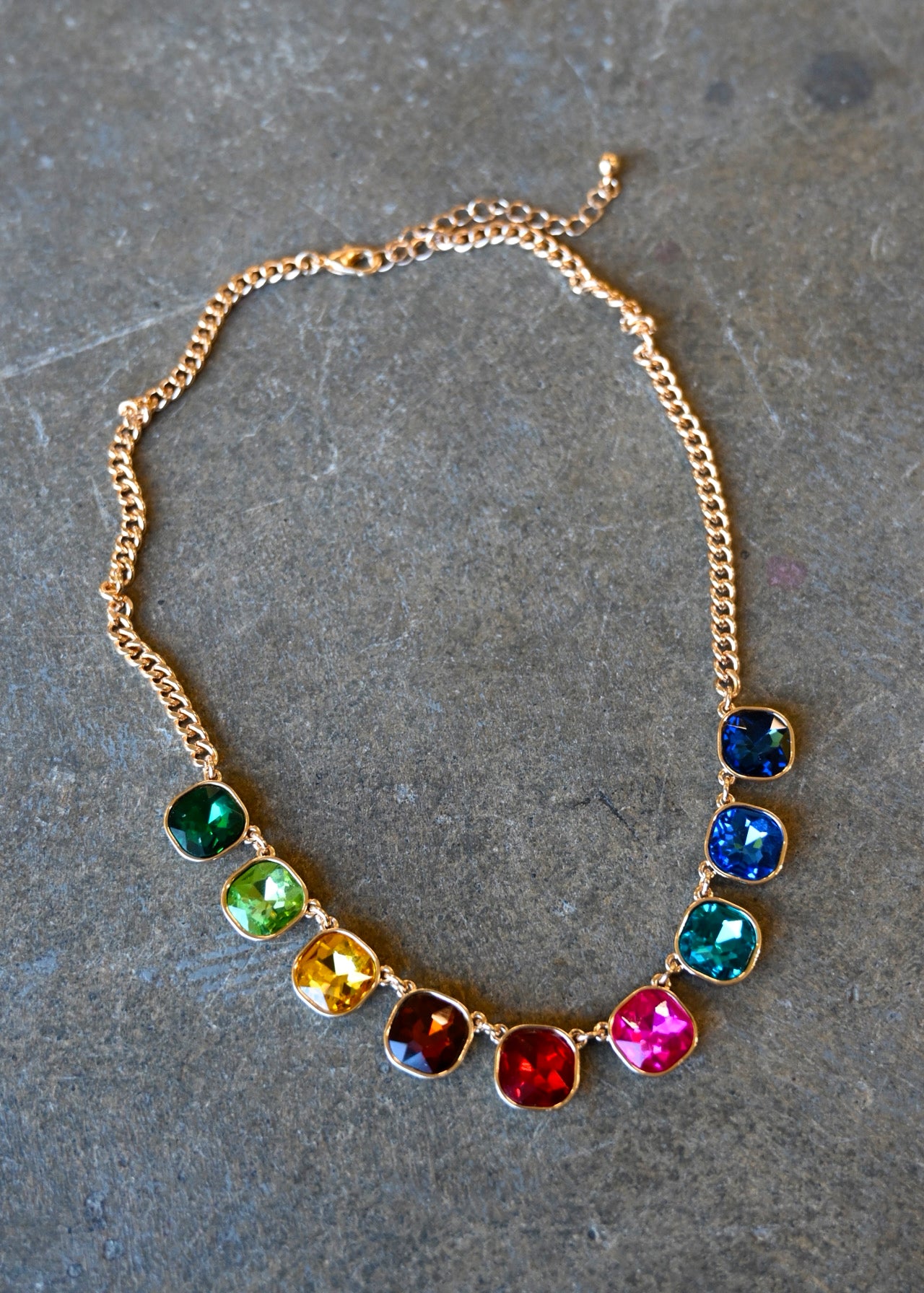 A gold necklace with multi-color cushion cut gemstones on a dark background
