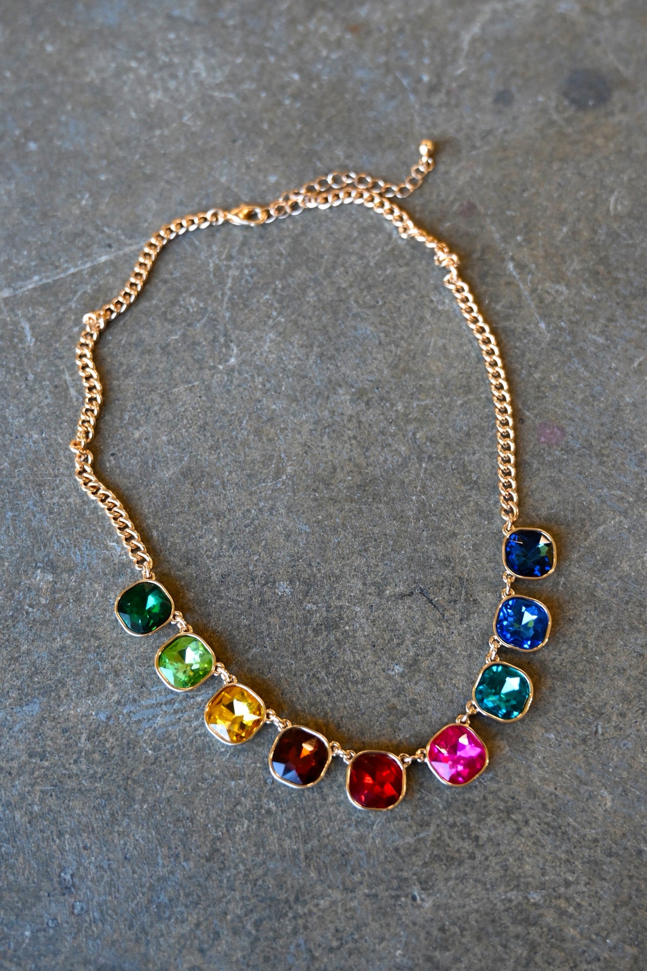 A gold necklace with multi-color cushion cut gemstones on a dark background