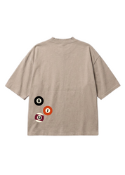 A beige short-sleeve t-shirt shown from the back, featuring a graphic of three billiard balls (black 8, orange 1, and red 13) on the lower left side, set against a plain white background