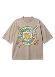 A beige short-sleeve t-shirt with a colorful graphic of a bowl of cereal on the front, featuring the text 'DON'T RUSH ME' and a spoon, set against a plain white background