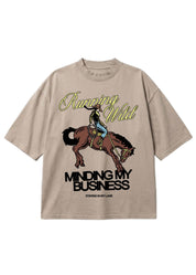 A beige graphic t-shirt featuring a cowboy riding a bucking horse, with the text 'Running Wild' in yellow script and 'MINDING MY BUSINESS' in black block letters. The brand 'STAYING IN MY LANE' is printed at the bottom of the graphic