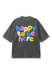 A dark gray washed short-sleeve t-shirt with a colorful graphic text print reading 'happy to be here' in a bubbly multicolor font with a yellow smiley face, shown on a plain white background.