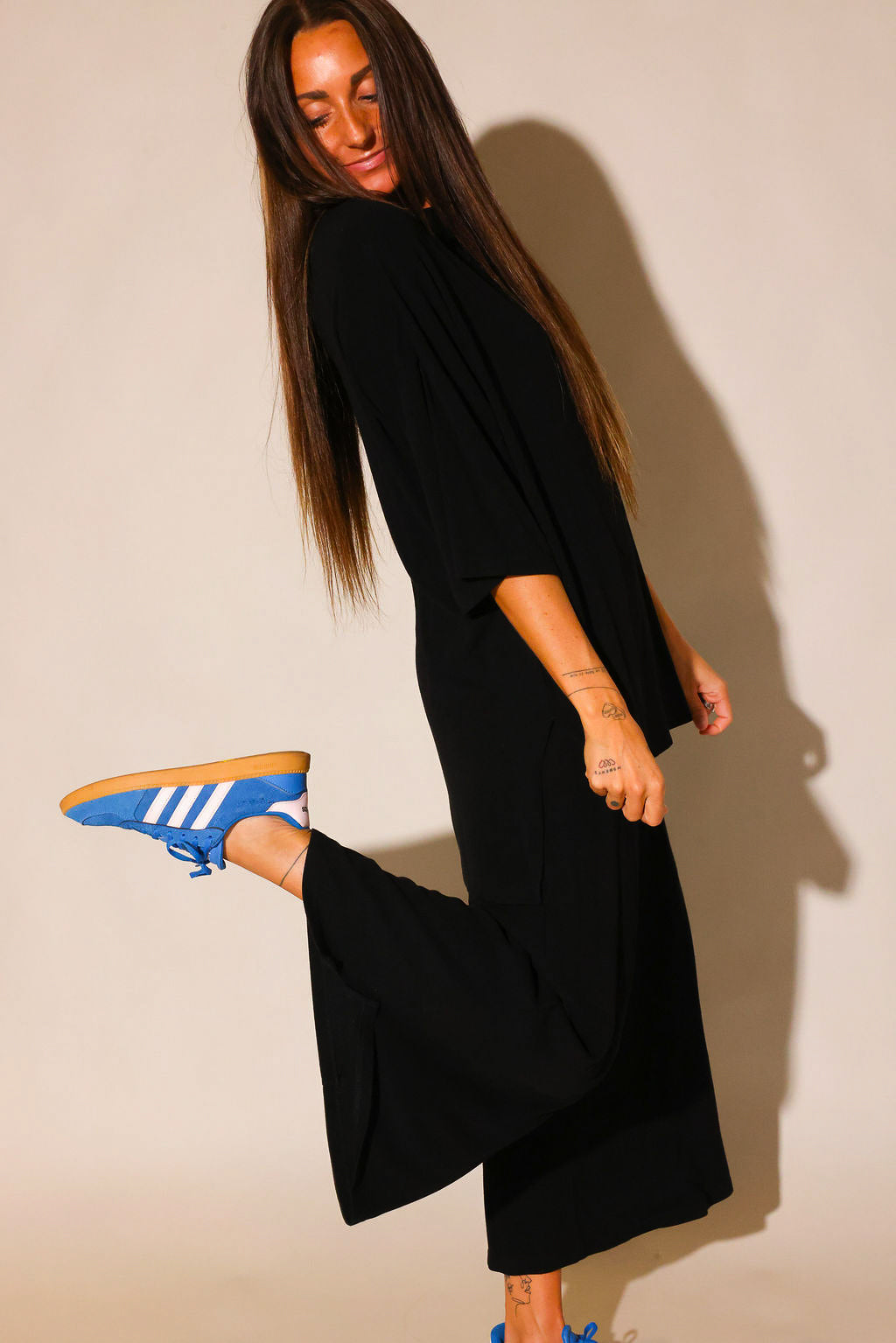 Side view of a model wearing a black ribbed short sleeve top and black capri pants, with blue Adidas sneakers, striking a playful pose
