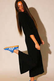Side view of a model wearing a black ribbed short sleeve top and black capri pants, with blue Adidas sneakers, striking a playful pose
