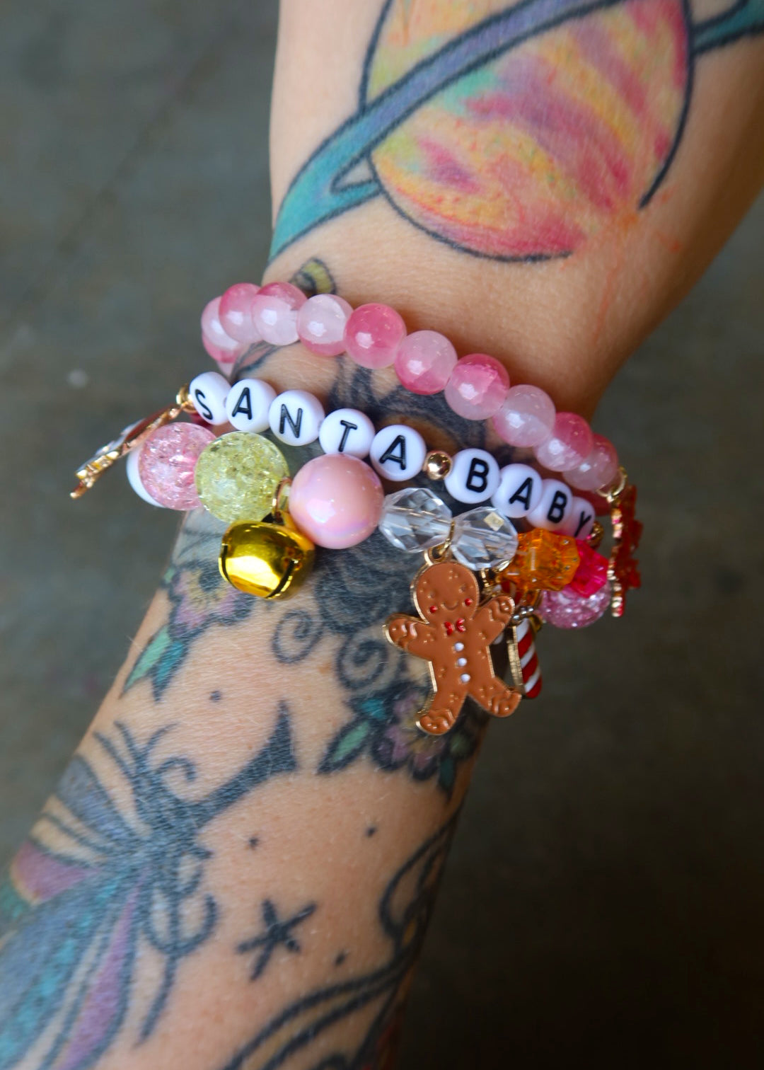Model wearing a stack of three bracelets with Christmas charms and beads that read 'Santa baby' on a dark surface