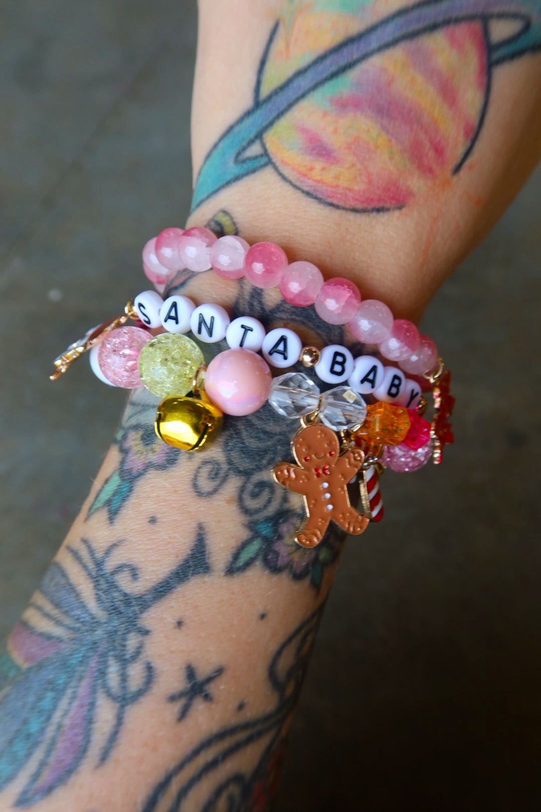 Model wearing a stack of three bracelets with Christmas charms and beads that read 'Santa baby' on a dark surface