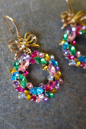 Close up of a pair of gold earrings with multicolored wreaths on a dark surface