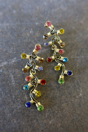 Pair of gold dangly earrings with a string of lights with multi-colored studs on a dark background