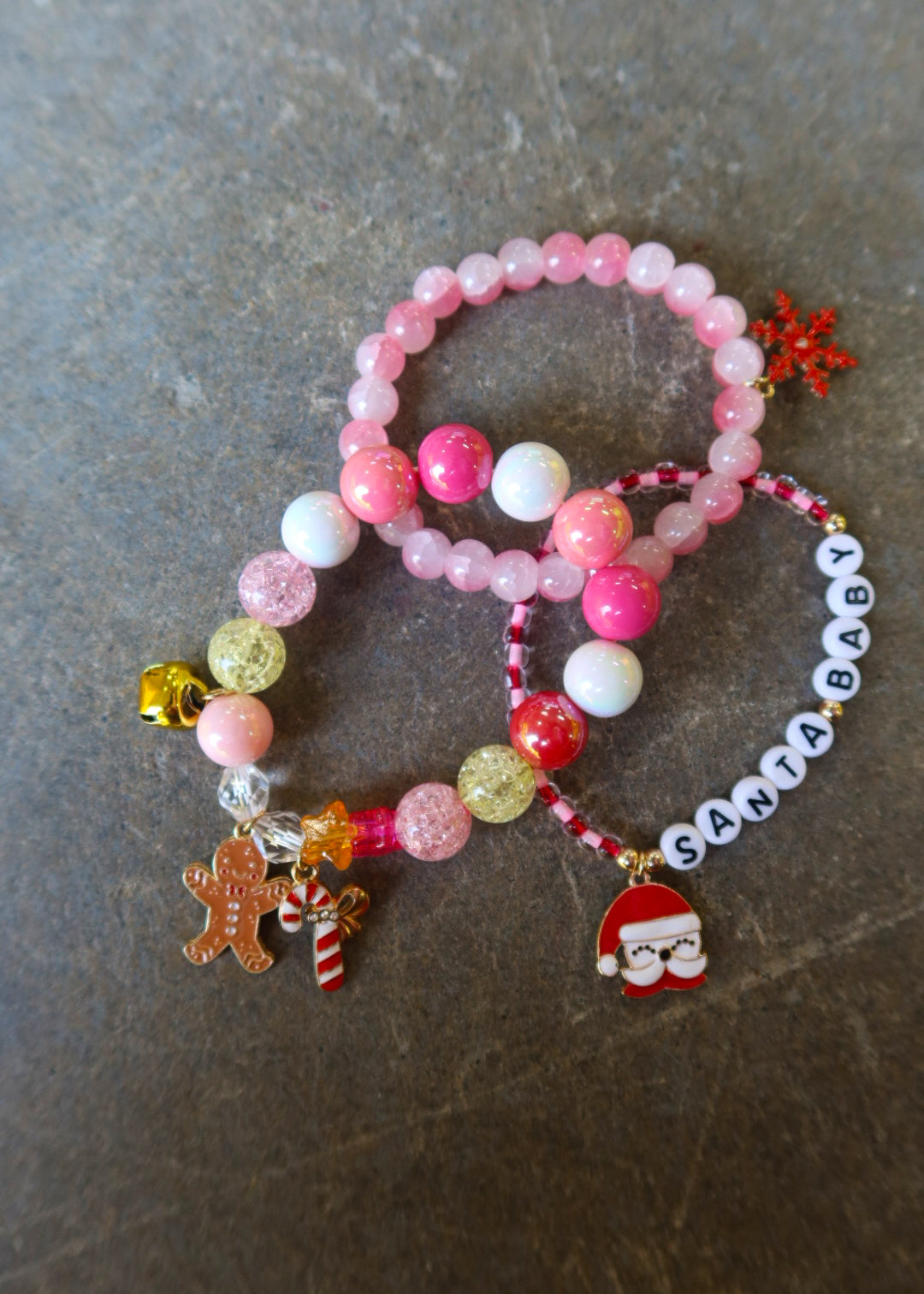 Stack of 3 bracelets with pink beads and Christmas charms and the words 'Santa baby' on a dark surface
