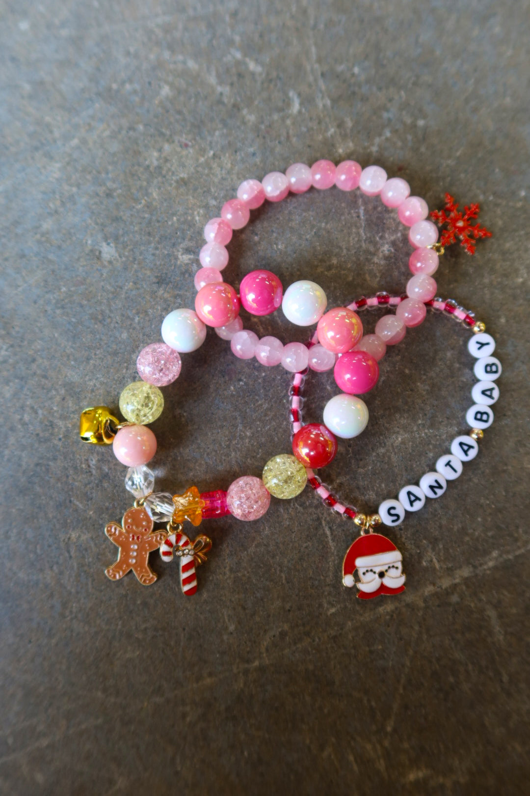 Stack of 3 bracelets with pink beads and Christmas charms and the words 'Santa baby' on a dark surface
