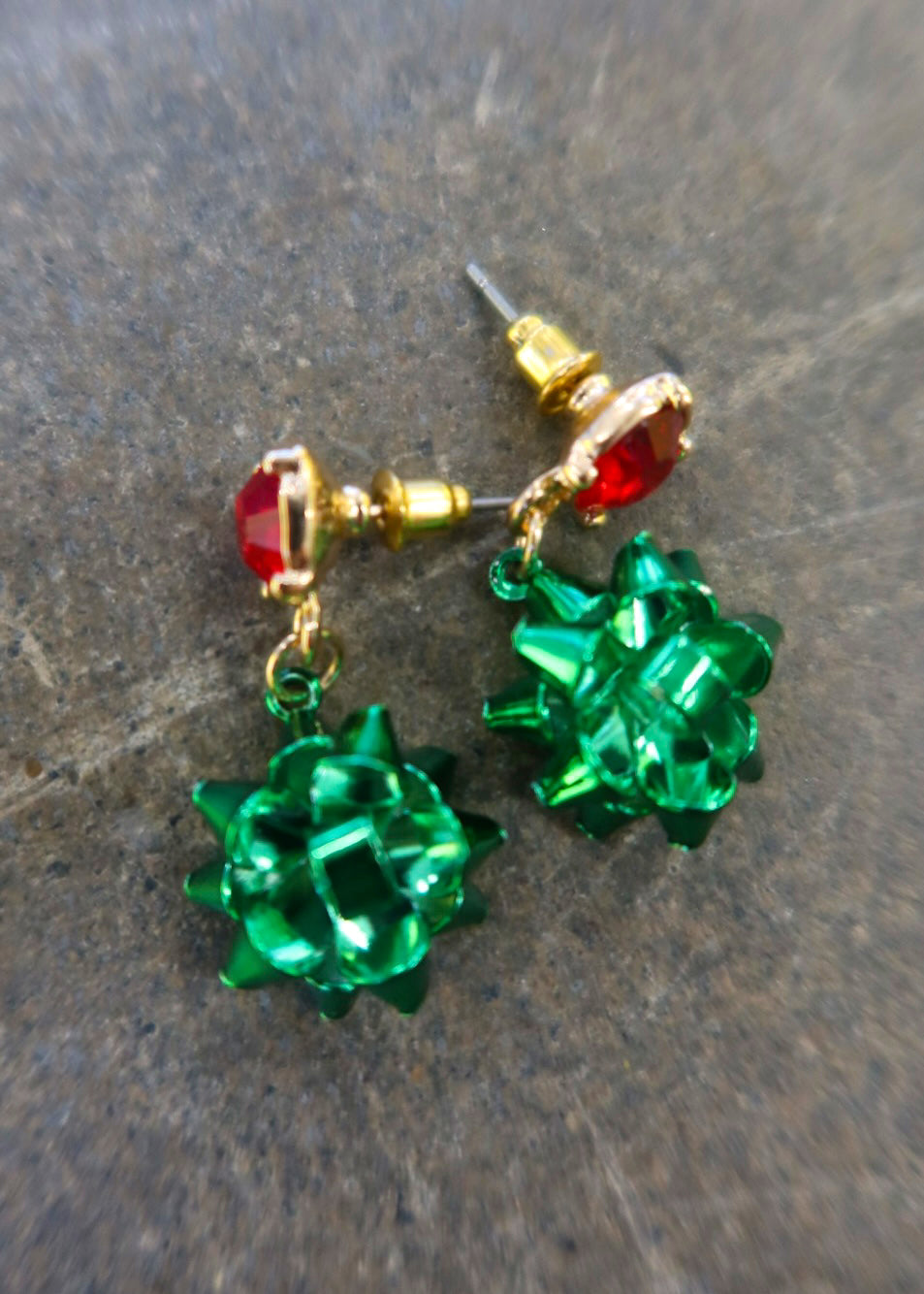 Gold dangly earring with a red stud and green bows on a dark background