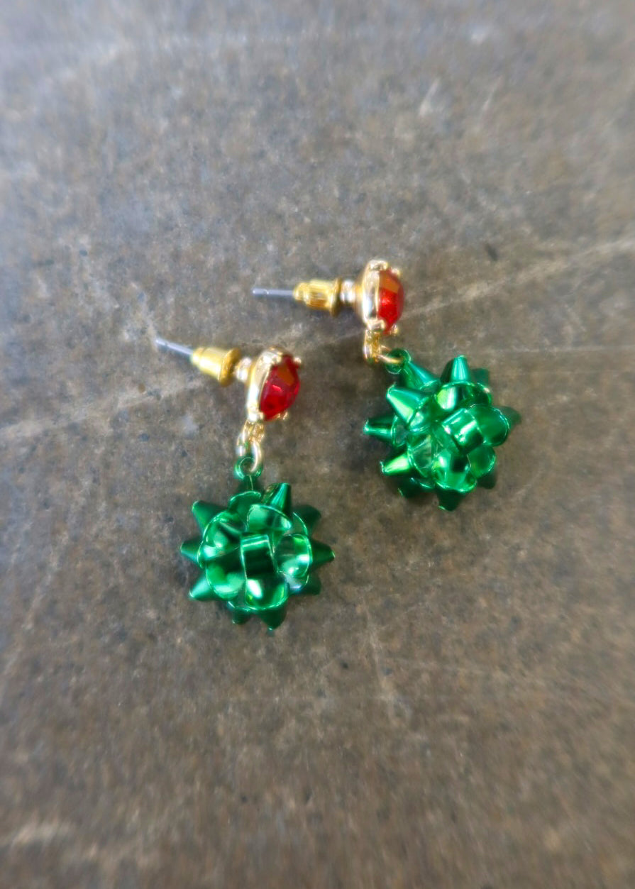 Gold dangly earring with a red stud and green bows on a dark background