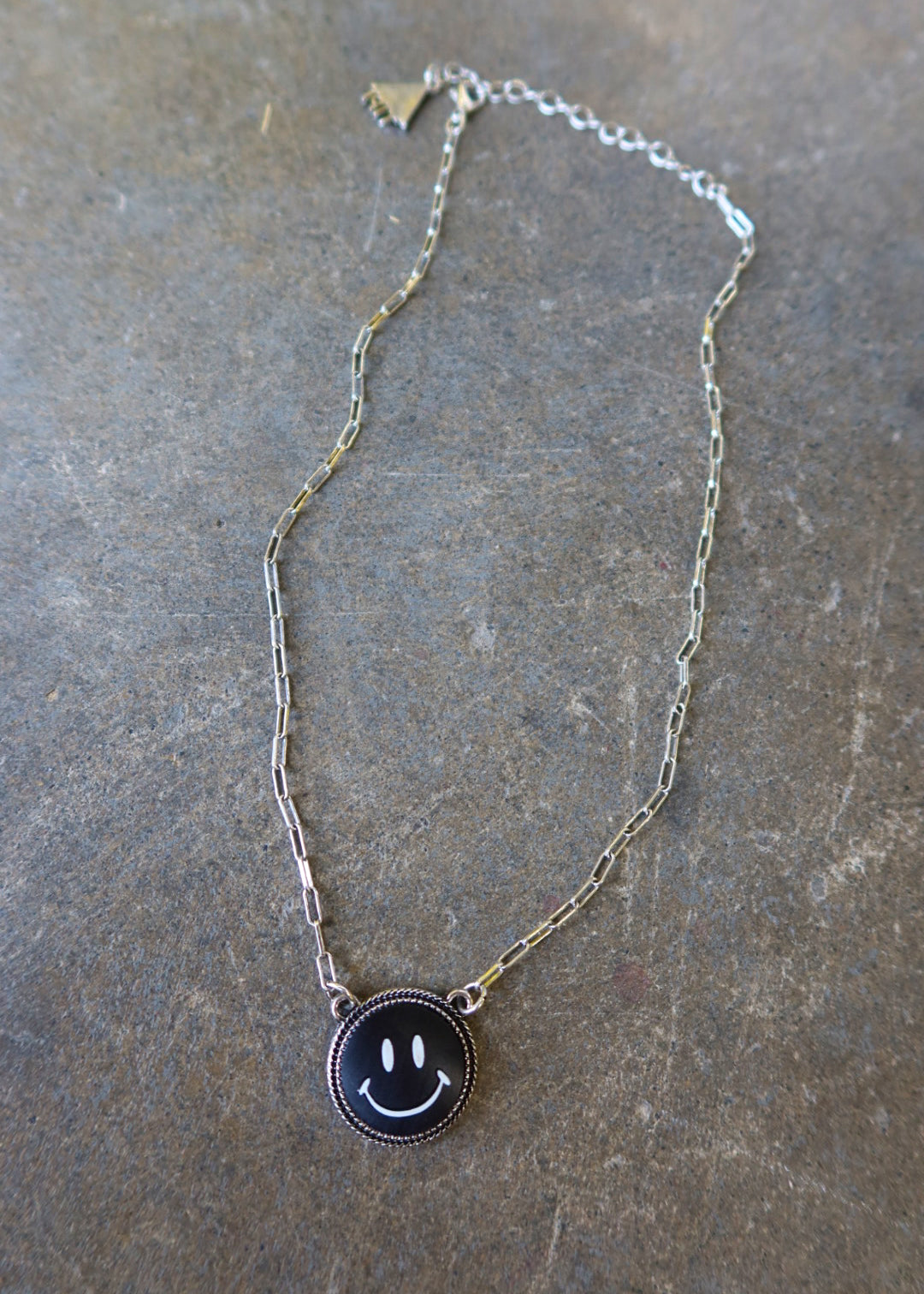 A silver necklace with a black smiley face charm on a dark surface