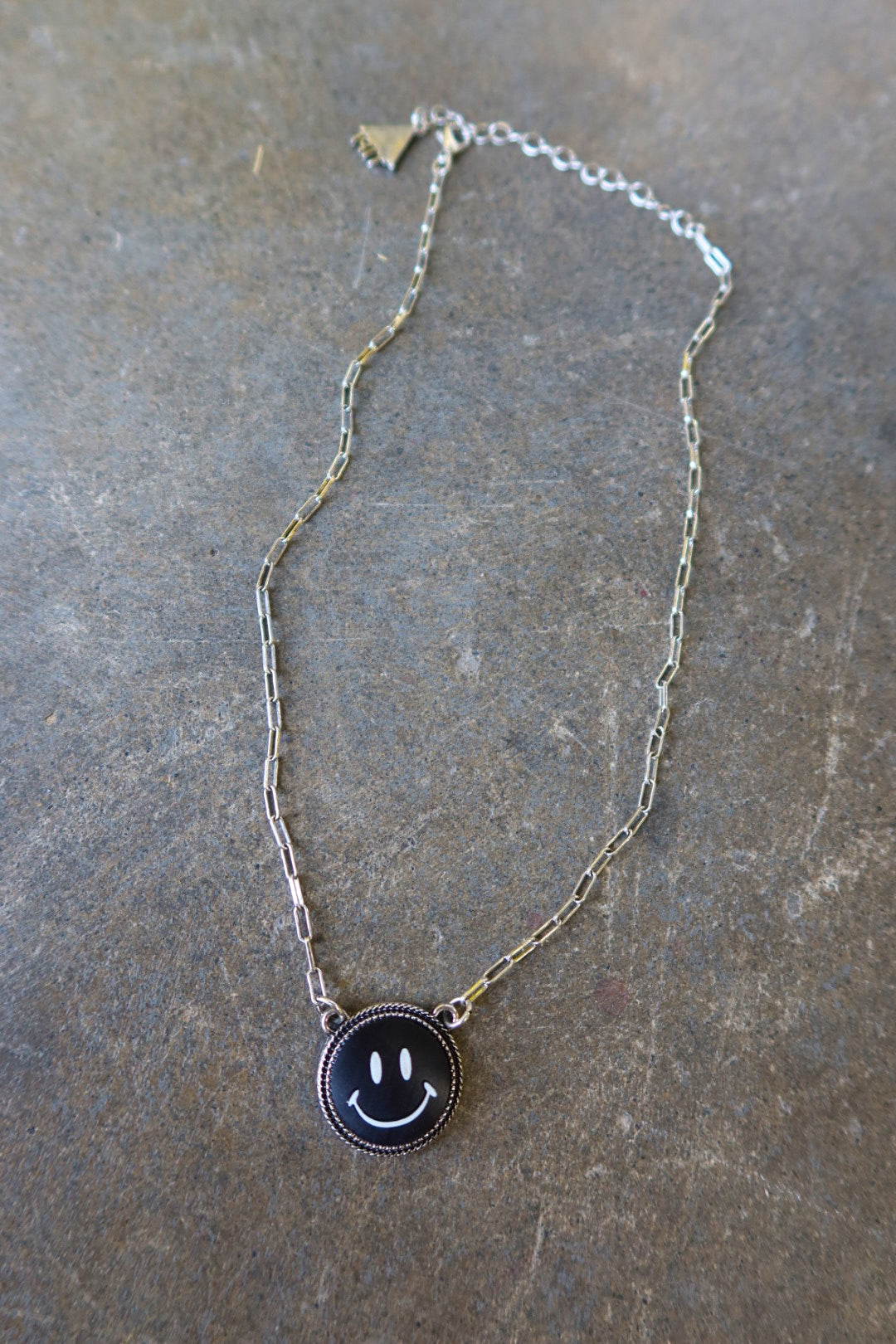 A silver necklace with a black smiley face charm on a dark surface