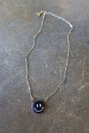 A silver necklace with a black smiley face charm on a dark surface