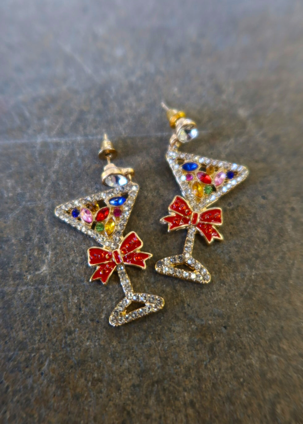 A pair of silver martini glass earrings with jewels and red bows on a dark background
