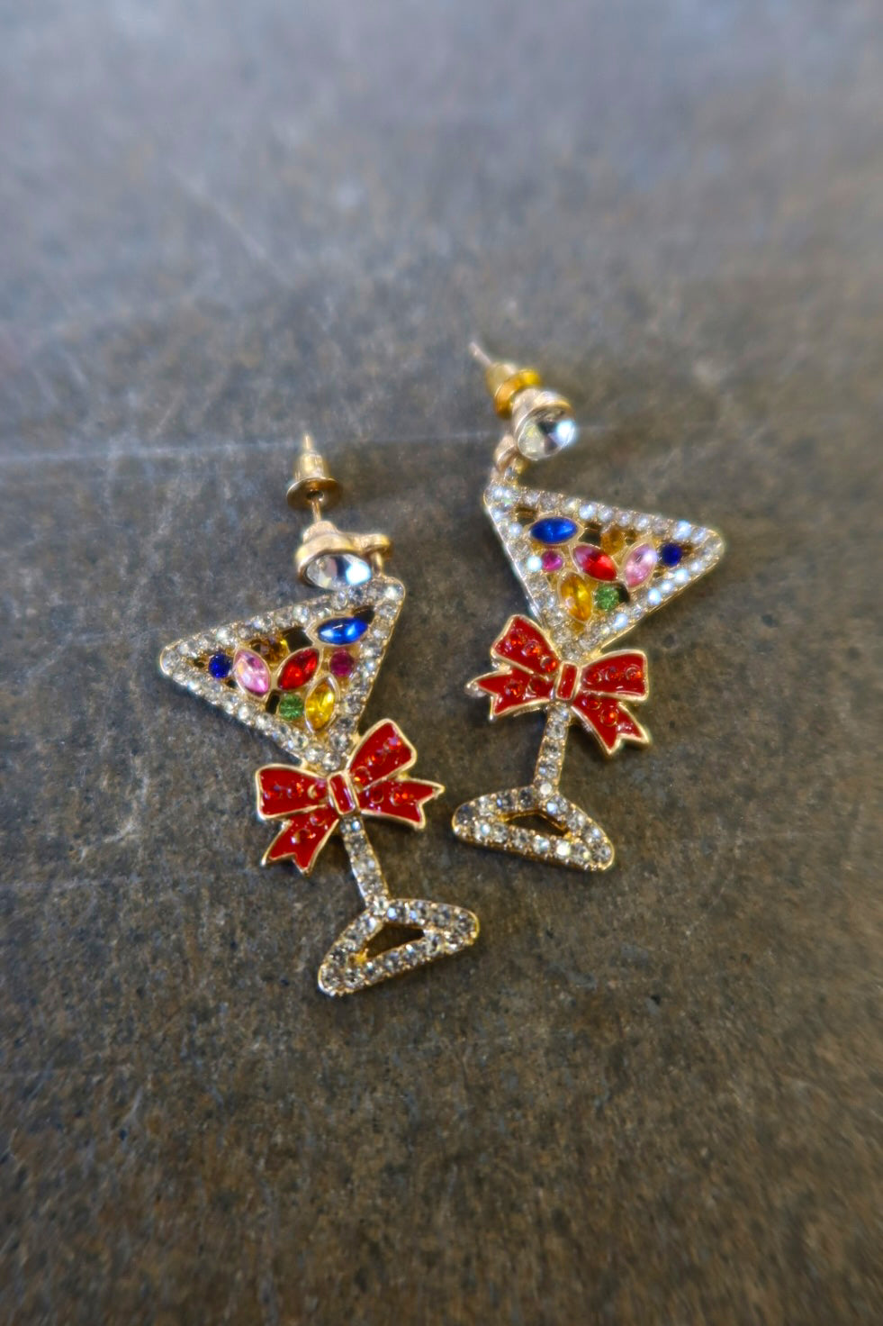 A pair of silver martini glass earrings with jewels and red bows on a dark background