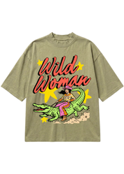 An oversized short-sleeve t-shirt in a washed olive green, featuring a retro graphic of a woman riding a green alligator with the text 'Wild Woman' in red script and yellow star accents