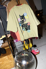 An oversized light olive green graphic T-shirt hanging on a rack, featuring the text 'bet on your self' in black with colorful numbered circles. The shirt is displayed in a clothing rack setup with other garments, a disco ball, and pink sneakers in a dim indoor setting.