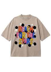 A beige short-sleeve t-shirt with a colorful graphic on the front that reads 'REACH FOR THE STARS' in multicolored letters, surrounded by black dots and white stars