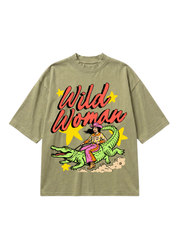 An oversized short-sleeve t-shirt in a washed olive green, featuring a retro graphic of a woman riding a green alligator with the text 'Wild Woman' in red script and yellow star accents