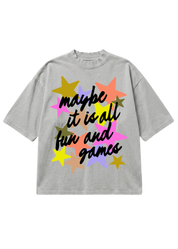 A light gray oversized short-sleeve t-shirt featuring a colorful graphic with the text 'maybe it is all fun and games' in black script over a cluster of pastel and bright stars
