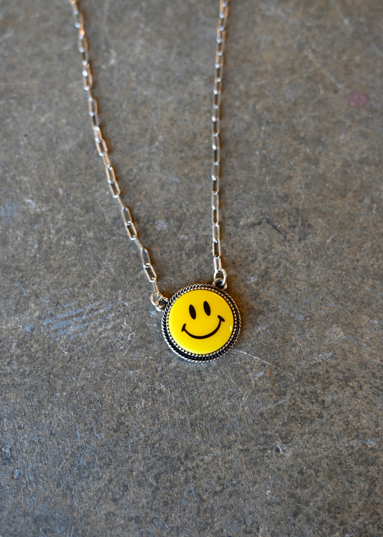 Close up of a silver chain necklace with a yellow smiley pendant on a dark background